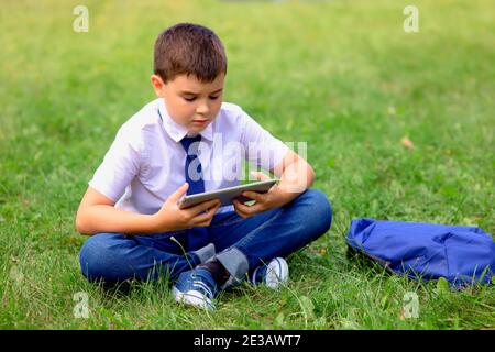 Un écolier sérieux est assis dans une chemise blanche et une cravate bleue sur l'herbe verte et regarde dans une tablette Banque D'Images