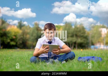 Un écolier heureux dans une chemise blanche s'assoit sur le vert herbe contre un ciel bleu avec des nuages blancs et tenir une tablette Banque D'Images