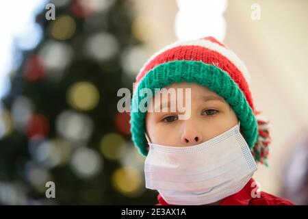 Un enfant dans un masque médical portant un chapeau du Père Noël en raison de la pandémie du coronavirus. Triste petit garçon portant un chapeau de Noël pendant le coronavirus epi Banque D'Images