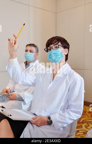 Jeune scientifique féminine en masque médical assistant à la conférence et à l'éducation main pour poser une question à l'orateur Banque D'Images