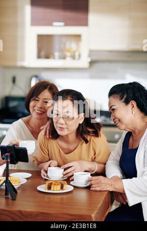 Souriantes femmes asiatiques qui boivent du thé avec des pâtisseries et des enregistrements vidéo pour leur chaîne Banque D'Images