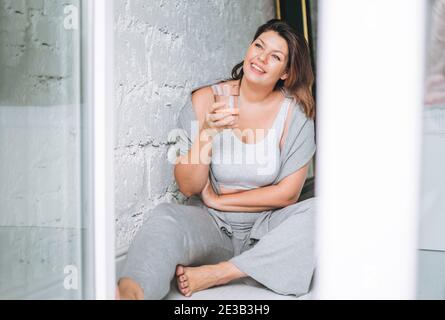 Belle heureuse affective brunette jeune femme plus taille corps positif dans un port de sport confortable avec un verre d'eau à la maison Banque D'Images