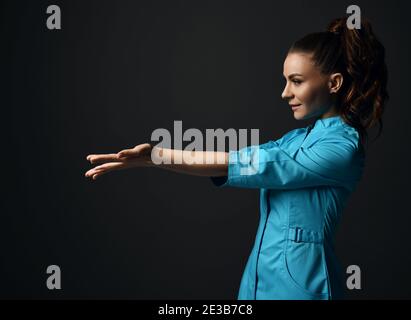 Jeune femme brune médecin infirmière en robe médicale bleue tient ses mains avec des paumes ouvertes avec l'espace de copie. Vue latérale Banque D'Images