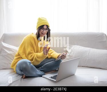 Élégante belle fille brune en lunettes et un chapeau assis sur le canapé devant un ordinateur portable et communique en ligne espace de copie. Banque D'Images