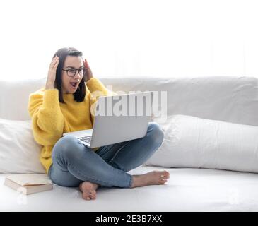 Une fille brune élégante dans un chandail jaune est assise à la maison sur le canapé et regarde avec surprise l'écran de l'ordinateur portable. Banque D'Images