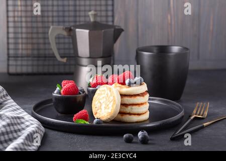 Crêpes au fromage cottage avec des framboises fraîches et des bleuets sur une assiette en céramique sombre. Tasse de café et cafetière - image Banque D'Images