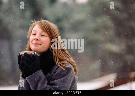 Une fille aux cheveux rouges satisfaite avec un nez rouge dans une forêt d'hiver se réchauffe avec du thé chaud parfumé. Scène d'hiver et émotions fortes. Banque D'Images