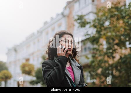 Belle femme pensive en particulier parlant au téléphone en marchant dans la rue avec une tasse de papier de café Banque D'Images