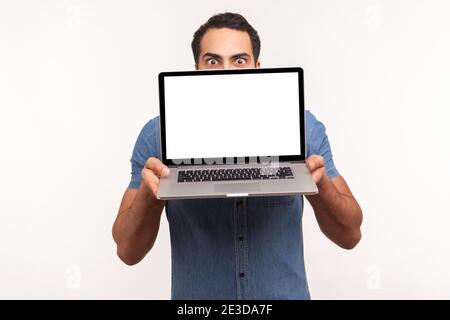 Homme en chemise bleue se cachant derrière blanc écran vide ordinateur portable, surpris par la zone de publicité, regardant la caméra avec de grands yeux, la mise à jour du système d'exploitation Banque D'Images