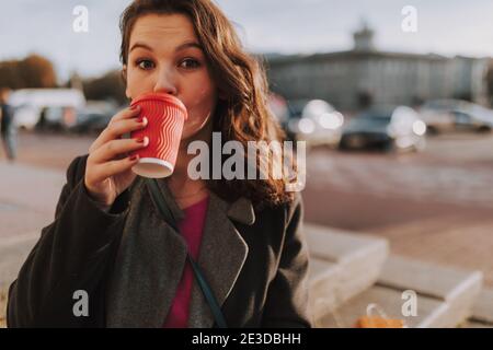 Gros plan de la jolie jeune femme debout dans la ville tout en dégustant un café avec des voitures en arrière-plan Banque D'Images