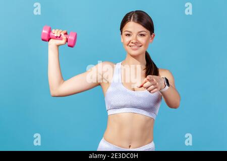 Femme sportive souriante en vêtements de sport blancs tenant la haltère rose dans la main et pointant le doigt vers l'appareil photo, vous recommandant de commencer à pomper les muscles. Banque D'Images