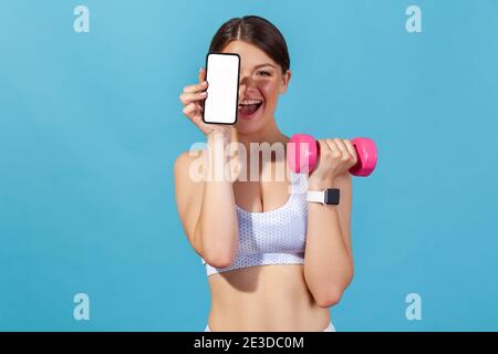 Happy Athletic femme en blanc sport top tenant un smartphone à écran vide et un dumbbell rose, faisant de la forme physique pilates s'exerçant regarder le tutoriel sur phon Banque D'Images