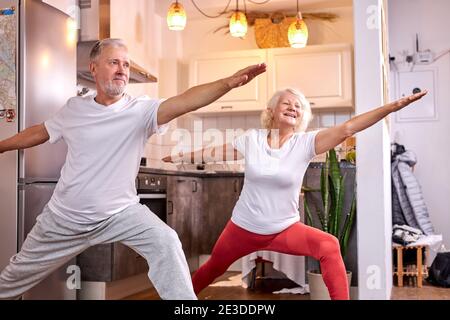 Homme et femme âgés pratiquant Virabhadrasana pose à la maison, yoga. Concept de mode de vie sain Banque D'Images