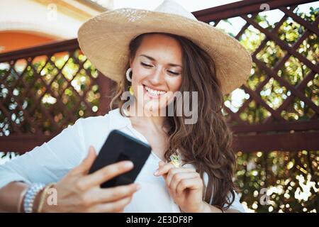 jeune femme souriante en chemise blanche avec chapeau ayant une réunion vidéo sur un smartphone sur la terrasse de l'hôtel de la maison d'hôtes. Banque D'Images