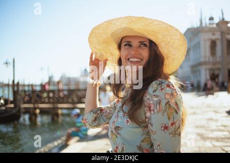 Femme de voyage moderne souriante d'âge moyen en robe florale avec chapeau ayant excursion sur la bankment à Venise, Italie. Banque D'Images