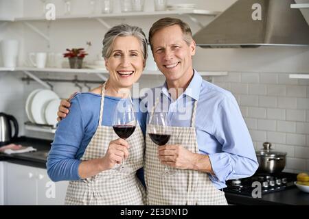 Joyeux vieux couple des années 50 qui s'embrasse en buvant du vin, regardant l'appareil photo dans la cuisine. Banque D'Images