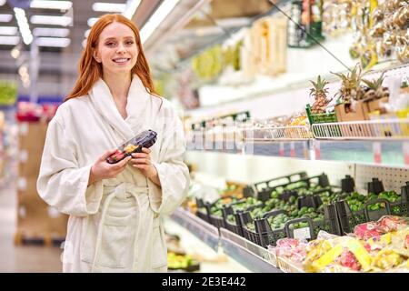 la femelle redhead tient les légumes emballés dans les mains, sourit à l'appareil photo, porte un peignoir. au supermarché Banque D'Images