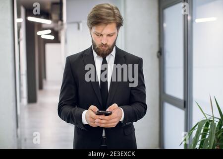 Homme d'affaires caucasien barbu concentré en tenue formelle, debout et utilisant le smartphone dans un bâtiment de bureau moderne à l'intérieur. Les gens, les affaires Banque D'Images