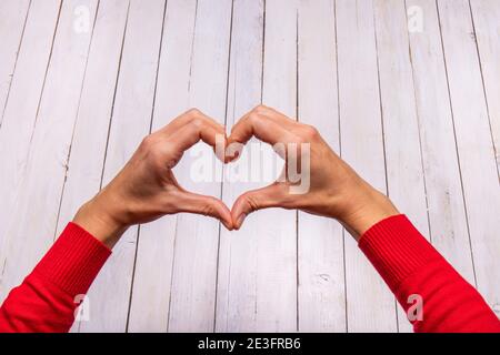 Les mains des femmes forment un cœur sur un fond en bois blanc. Une touche de rouge du pull attire l'attention. Espace vide pour le texte de la Saint-Valentin. Banque D'Images