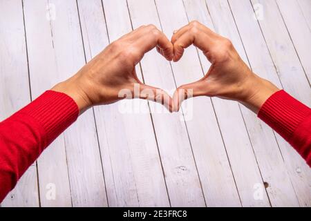 Les mains des femmes forment un cœur sur un fond en bois blanc. Une touche de rouge du pull attire l'attention. Espace vide pour le texte de la Saint-Valentin. Banque D'Images