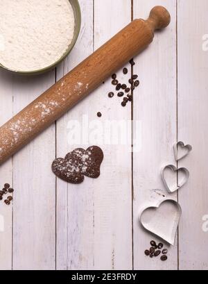 Faire des biscuits pour la période la plus douce de l'année. Deux biscuits au chocolat avec un bol de farine et une broche sur une table en bois blanc avec un peu de café Banque D'Images