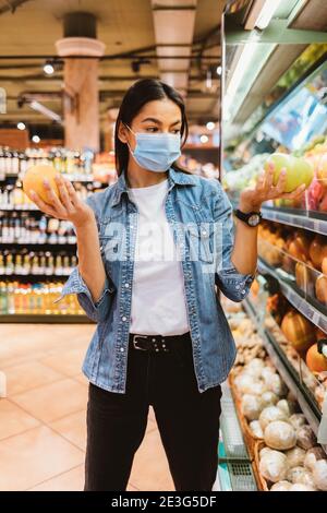 Une jeune femme en masque achète des oranges dans le magasin, image verticale Banque D'Images