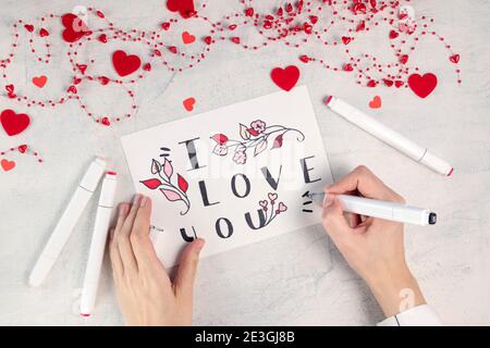 Les mains des femmes font la carte de Saint-Valentin avec l'inscription Je t'aime sur la table blanche avec des perles rouges. Préparation de la Saint-Valentin. Concept de la Saint-Valentin. Pose à plat. Vue de dessus Banque D'Images