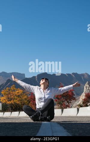 La dame en sweat à capuche blanc et le Jean est assis sur l'asphalte gris route qui s'étend les bras avec joie contre les arbres colorés et rocailleux montagnes aux rayons du soleil Banque D'Images