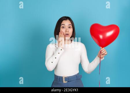 Surprise, choquée jeune femme asiatique à la bouche ouverte tient un ballon volant en forme de coeur isolé sur un fond bleu Banque D'Images