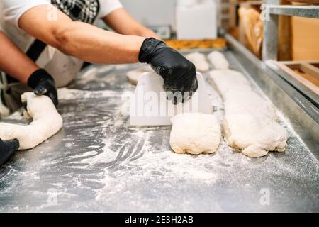 Mise au point sélective de deux boulangers latins pétrissant et coupant le pétrissez pour faire du pain avant de le mettre au four sur une table de cuisson avec des gants noirs Banque D'Images