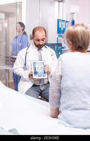 Médecin expliquant une lésion cérébrale à une femme âgée sur un Tablet pc dans la salle d'examen de l'hôpital. Thérapeute médical montrant et parlant de la radiographie avec le patient pendant la consultation. Banque D'Images