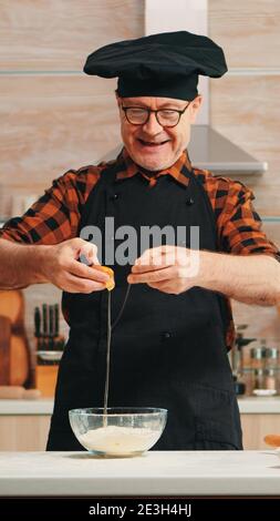 Un boulanger expérimenté craque des œufs pour cuire portant un tablier en appréciant le passe-temps. Ancien chef de cuisine avec mélange bonete à la main, pétrissage dans un bol en verre ingrédients de pâtisserie préparation de gâteau maison Banque D'Images