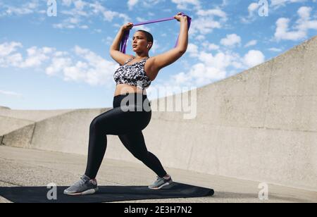 Pleine longueur de femme de taille plus s'exerçant avec la bande de résistance. Une femme fait de l'exercice avec un bracelet de résistance à l'extérieur le matin. Banque D'Images