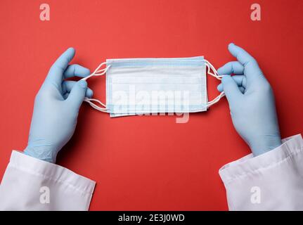 deux mains de sexe masculin dans des gants en latex bleu tiennent un médecin masque jetable sur fond rouge Banque D'Images