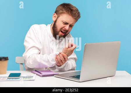 Homme barbu employé de bureau en chemise blanche grimacing sensation de picotements, engourdissement et douleur au poignet travaillant sur ordinateur portable, souffrant du syndrome du tunnel carpien. JE Banque D'Images