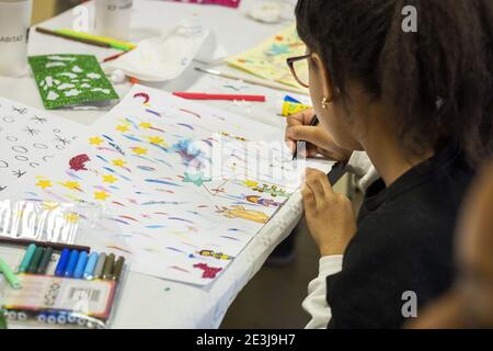 Concours de dessin de Noël pour enfants. Vue de trois quarts arrière d'une fille portant des lunettes et des boucles d'oreilles dorées, dessinant une série de personnages avec un Banque D'Images