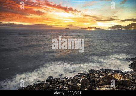 Vue panoramique unique et longue exposition sur le récif et une mer agitée contre le ciel orange du feu et le village de Portovenere, Tellaro, la Spezia, Italie Banque D'Images