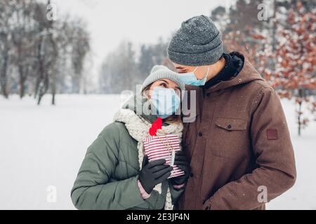 L'homme donne une boîte cadeau à sa petite amie pour la Saint Valentin dans un parc d'hiver enneigé. Le couple porte des masques contre les mesures de sécurité en cas de pandémie de covid du coronavirus Banque D'Images