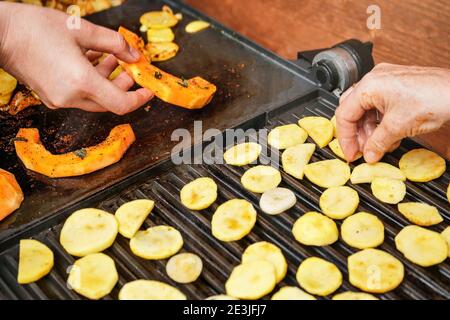 Morceaux de courge de noyer cendré orange grillés sur le gril électrique, détail sur les mains de femme en train de fumer des légumes, chips de pomme de terre floues à l'avant Banque D'Images