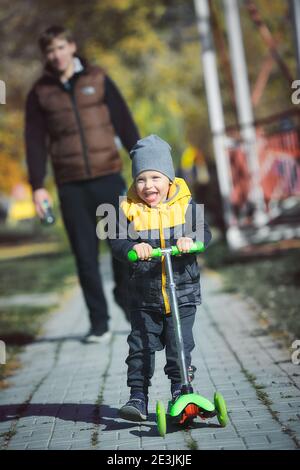 Un petit garçon heureux sur un scooter marche dans le parc avec son père un jour d'automne. Banque D'Images