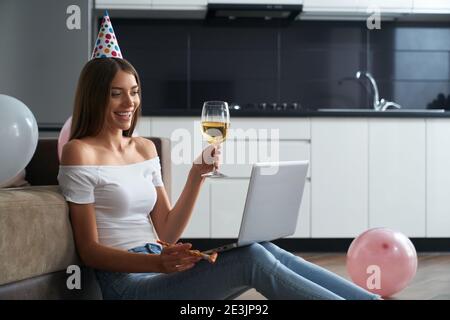 Jeune femme joyeuse ayant une vidéoconférence sur un ordinateur portable moderne tout en étant assise sur le sol avec un verre de vin. Femme souriant, dorant et célébrant birt Banque D'Images