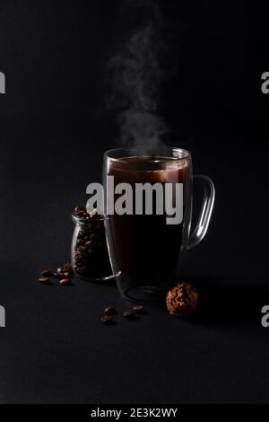 Café noir chaud dans une tasse de verre avec truffe de chocolat et beaucoup de grains de café placés dans un pot et autour sur fond sombre. Vue avant Banque D'Images