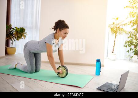 Jeune femme en forme effectuant des exercices d'abs via un ordinateur portable à l'aide d'un rouleau de gymnastique. Banque D'Images