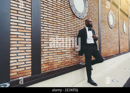 Un élégant homme africain à barbe chauve mature dans un noir costume avec une chemise blanche et un noeud papillon est penché contre un mur moderne en brique avec le vent de hublot Banque D'Images