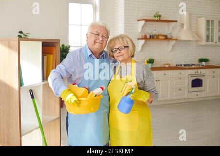 Couple de personnes âgées debout dans la cuisine dans des tabliers tenant des détergents pour nettoyer la maison. Banque D'Images
