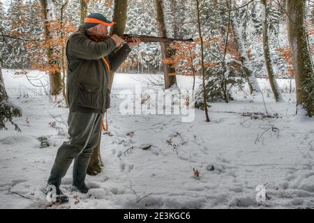 Un ancien chasseur expérimenté se tient près d'un arbre dans la forêt noire enneigée et tient sa carabine de chasse en place. Banque D'Images