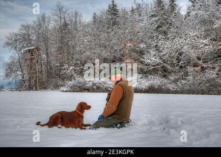 Dans la forêt noire enneigée, un chasseur s'agenouille dans la neige devant sa grande chaire de chasse, devant lui se trouve sa belle chasse au Setter irlandais Banque D'Images