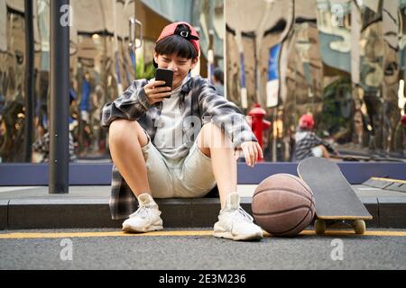 adolescent asiatique avec basketball et planche à roulettes assis sur le trottoir de la rue donnant sur le téléphone portable Banque D'Images