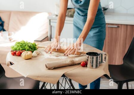 Femme femme au foyer dans un tablier dans la cuisine roule la pâte à pizza. Banque D'Images