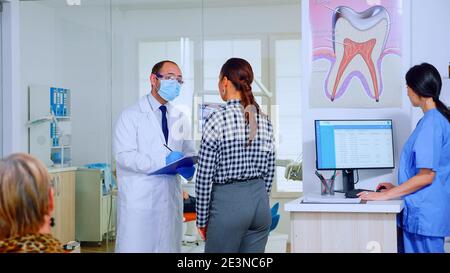 Dentiste médecin interrogeant la femme et prenant des notes sur le presse-papiers debout dans la salle d'attente. Jeune patient expliquant le problème dentaire au stomatologiste parlant dans la réception surpeuplée de la clinique. Banque D'Images